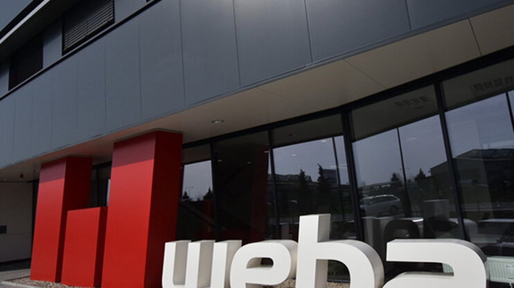 Fotoaufnahme vor dem Eingang von weba Olomouc: Anthrazitfarbene Gebäudefront mit Fensterfront, davor das weba-Logo in Rot und Weiß in großen 3D-Buchstaben.