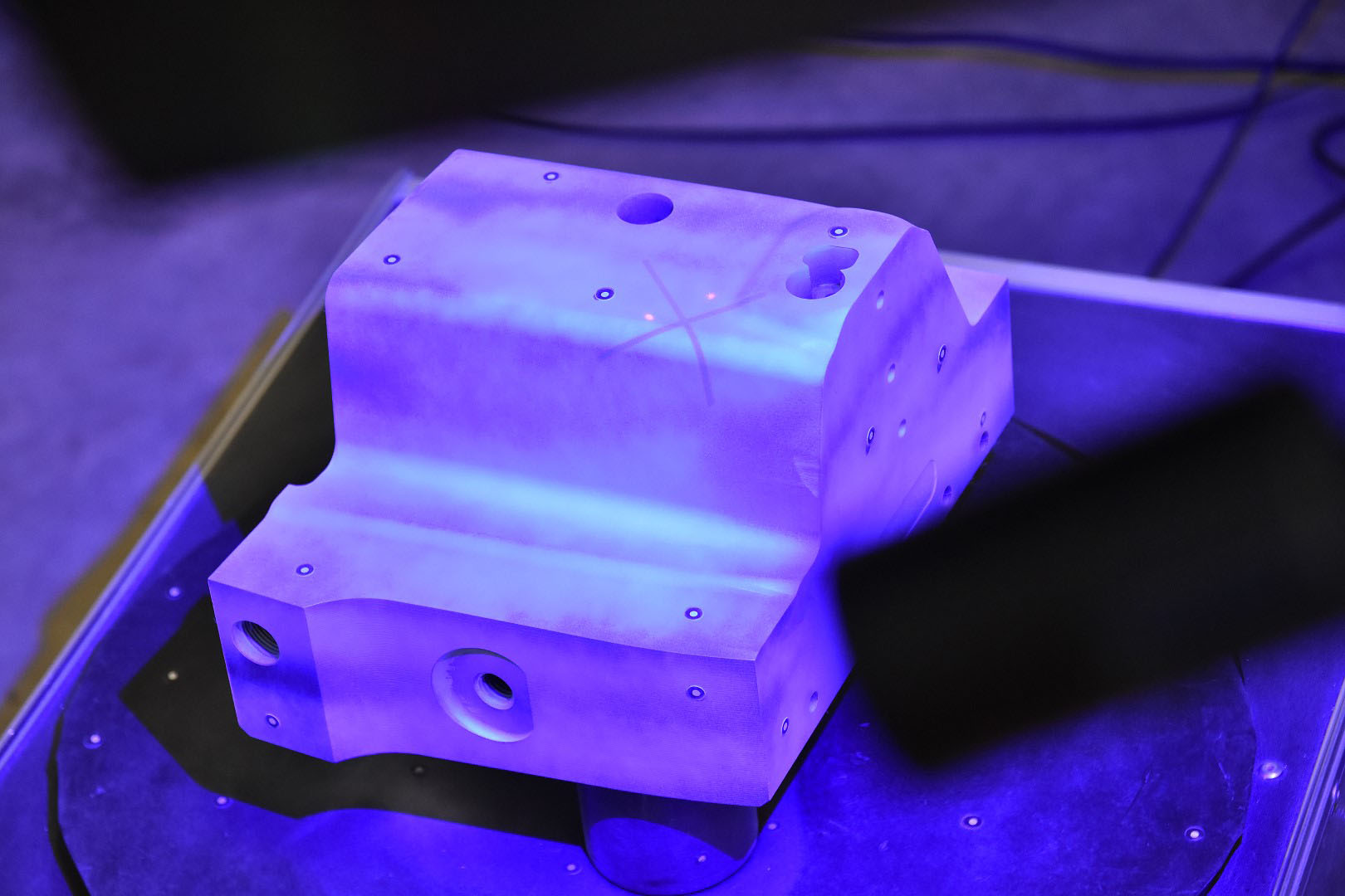 Ein Stahleinsatz befindet sich zentriert auf einer Drehscheibe in einem technischen Labor. Ein 3D-Scanner, der über der Drehscheibe positioniert ist, digitalisiert und analysiert das Objekt sorgfältig.