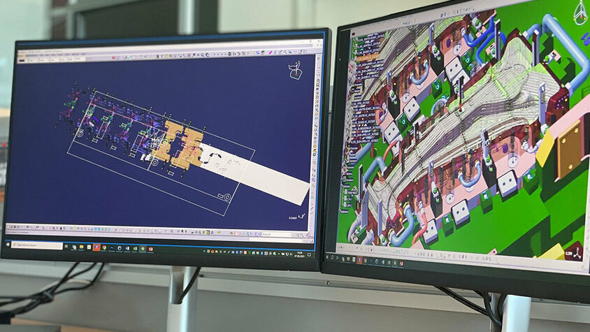 Zwei PC-Bildschirme, einer zeigt ein detailliertes Close-up einer CAD Konstruktion eines Warmumformwerkzeuges zur Produktion von B-Säulen, während der andere Bildschirm den Transferaufbau eines Werkzeuges darstellt.