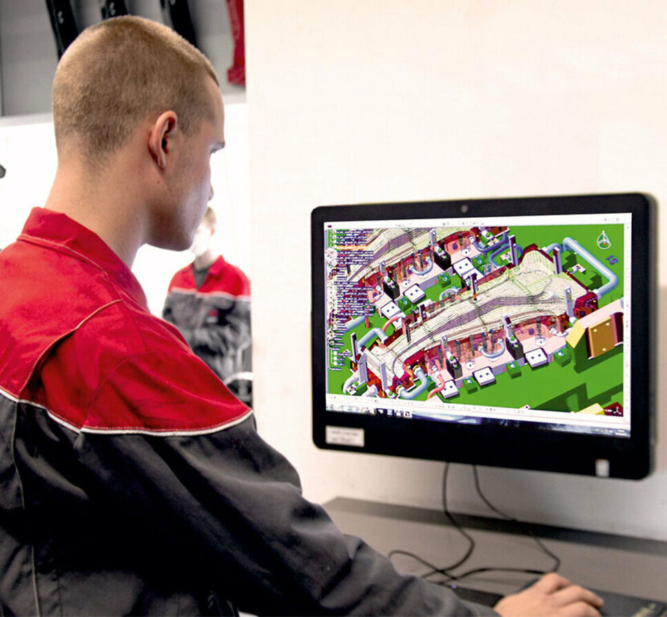 An apprentice in toolmaking/metal technology is looking carefully at a 3D design of a hot forming tool on a PC screen. He is studying the details and structure of the tool in preparation for the upcoming tool assembly. His focused posture shows how he is grasping the design and complexity of the tool in order to mentally prepare himself for the practical implementation of the design in his metal technology apprenticeship.