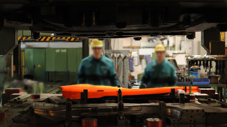 Glühende Stahlplatine in Umformwerkzeug mit Produktion im Hintergrund und verschwommenen Werkzeugbautechnikern