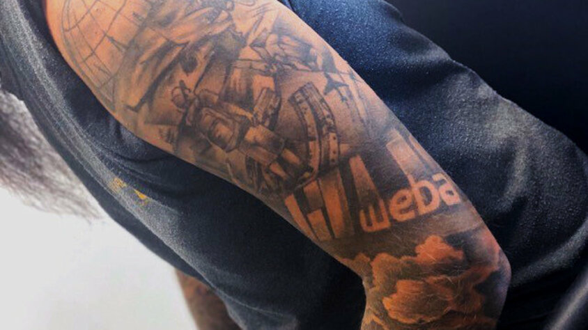 Ein farblich dezentes Oberarmtattoo eines Mitarbeiters, das das Logo des Unternehmens „weba“ zeigt. Um das Logo herum sind detaillierte technische Elemente wie Zahnräder, Maschinen und eine Weltkugel abgebildet. Das Tattoo symbolisiert die enge Verbindung des Mitarbeiters zu weba Werkzeugbau und seiner Leidenschaft für Technologie und Präzision.