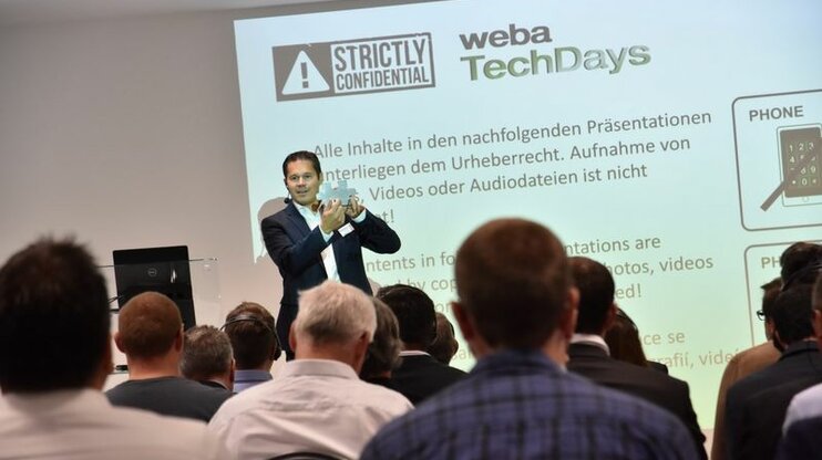 Geschäftsführer Hannes Feuerhuber von weba Werkzeugbau begrüßt das Publikum der Werkzeugbaukonferenz auf der Bühne, während im Hintergrund die Geheimhaltungsrichtlinien projiziert werden.