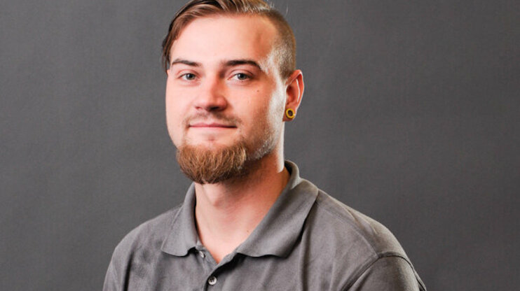 Porträtfoto von Bronislav Škoda, einem jungen Mann mit kurzem, seitlich rasiertem Haar und einem gepflegten Vollbart. Er trägt ein graues Poloshirt mit dem Logo von weba und schaut freundlich in die Kamera. Seine Ohren sind mit gelben Plugs geschmückt, und im Hintergrund ist eine neutrale graue Studioleinwand zu sehen.