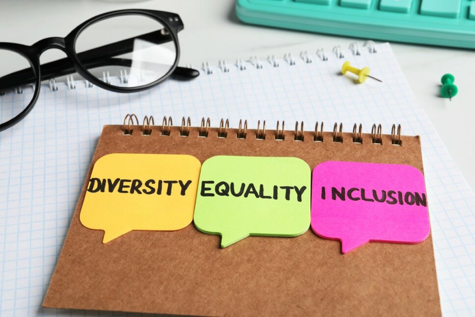 Hinterseite eines Blocks mit drei bunten Post-its, auf denen die Wörter 'Diversity, Equality und Inclusion' stehen. Daneben liegen eine schwarze Rahmenbrille, eine türkisfarbene Tastatur sowie zwei Pinnadeln in grün und gelb.