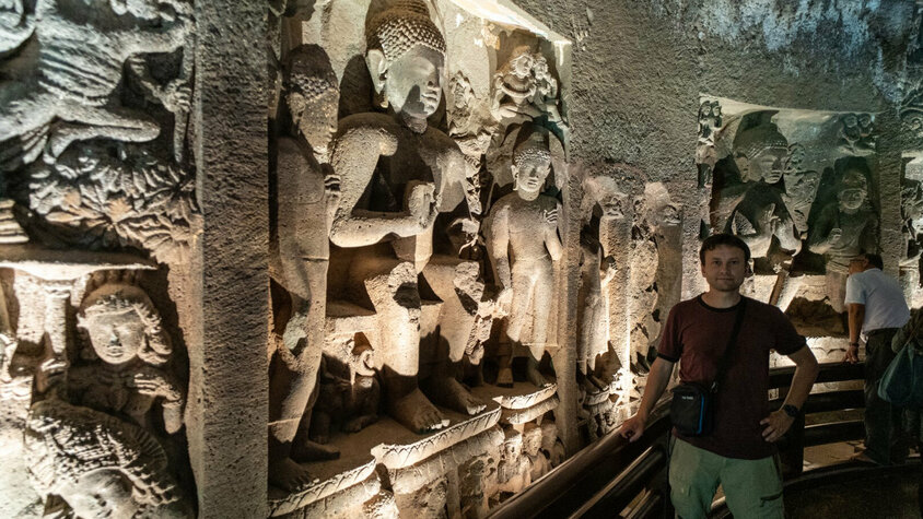 Eine Innenansicht einer Höhle mit kunstvoll gemeißelten Reliefs, die Buddha und andere Figuren darstellen, beleuchtet von weichem Licht. Eine Person steht im Vordergrund und betrachtet die beeindruckenden Schnitzereien.