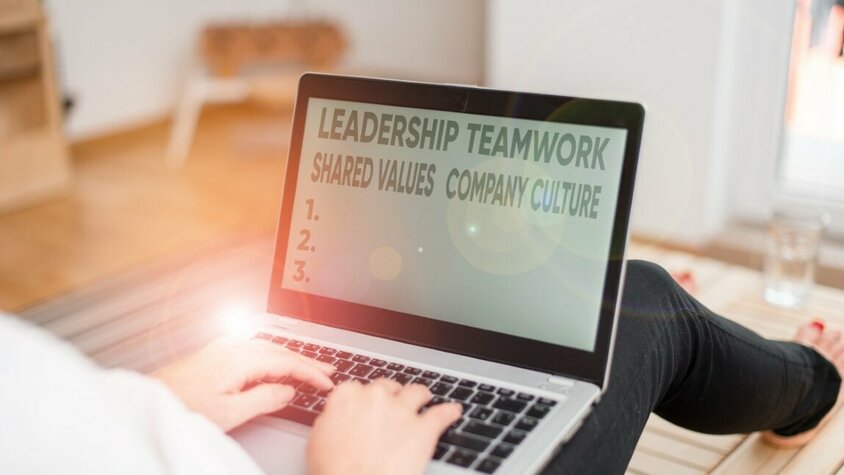 Eine Frau sitzt auf dem Boden mit einem Laptop auf ihrem Schoß. Auf dem Bildschirm sind die Wörter 'Leadership, Teamwork, Shared Values und Company Culture' zu lesen, während das Licht auf dem Laptop reflektiert wird. Das Bild symbolisiert wertegebundene Arbeitskultur bei weba für deine Karriere in Steyr.