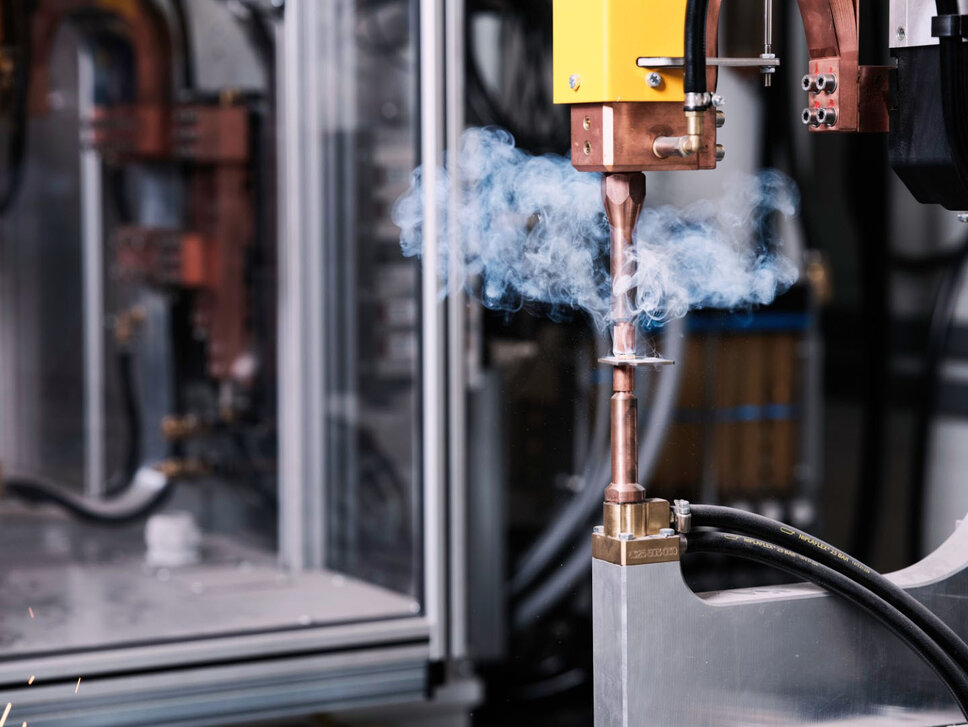 Close-up of an industrial welding system in a welding lab. The machine performs precise spot welding as smoke rises. In the background, complex technical equipment and lab facilities used for advanced welding processes can be seen.