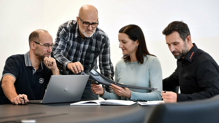 Vier Personen sitzen an einem Besprechungstisch: Eine Person arbeitet an einem Laptop, eine andere betrachtet aufmerksam Unterlagen, während zwei weitere Personen sich angeregt über ein Stanzteil unterhalten. Die Szene zeigt eine konzentrierte und engagierte Arbeitsatmosphäre. Berufserfahrene und Profis finden bei weba ihren Traumjob in Steyr. 