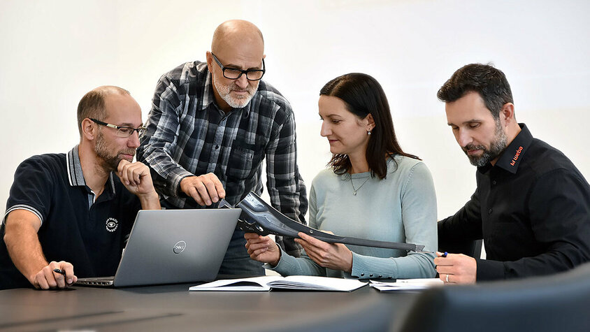 Vier Personen sitzen an einem Besprechungstisch: Eine Person arbeitet an einem Laptop, eine andere betrachtet aufmerksam Unterlagen, während zwei weitere Personen sich angeregt über ein Stanzteil unterhalten. Die Szene zeigt eine konzentrierte und engagierte Arbeitsatmosphäre. Berufserfahrene und Profis finden bei weba ihren Traumjob in Steyr. 