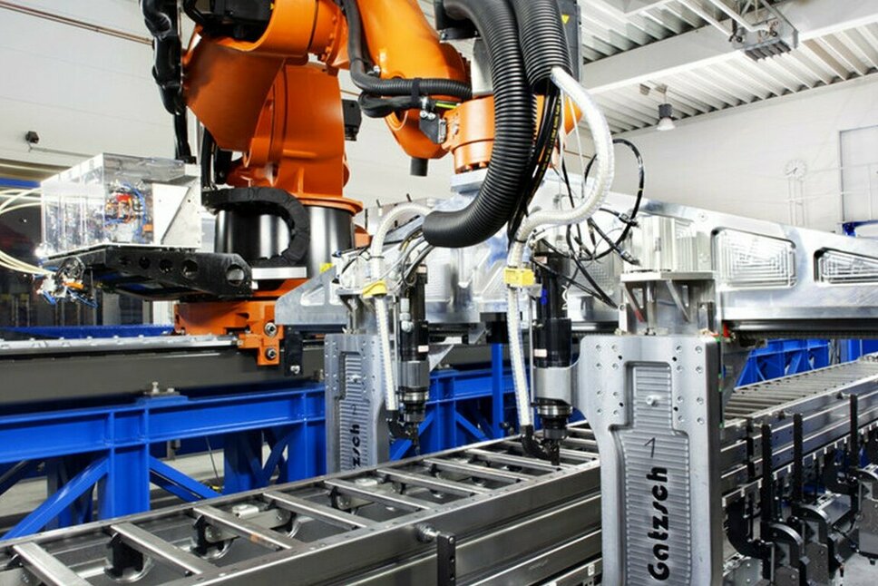 A modern industrial robot working in a production hall at an automated manufacturing line. The robot is performing riveting and clinching tasks to precisely join metal components. The system is attached to a conveyor and is situated in a bright, industrial environment, representing efficient and precise processes in joining technology.