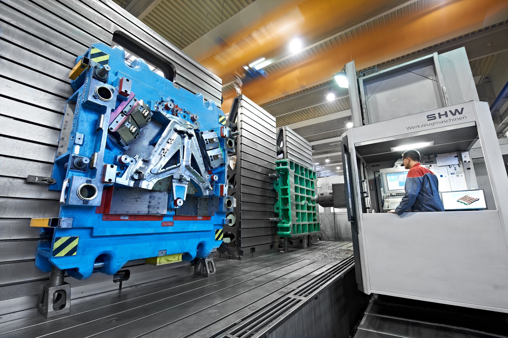 A technician operates a large SHW machine tool in a production hall. On the left, a large blue component is fixed in place, ready for machining. In the background, additional components and machine parts are visible.